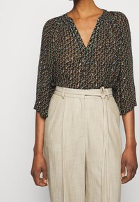 Svart blommig blus med V-ringning, långa ärmar och rynkade axlar; tillsammans med beiga högmidjade byxor med bälte.