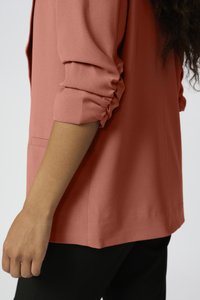 Korálová blůza s ohrnutými rukávy, jemná struktura tkaniny, detail boční kapsy, nošena přes černé kalhoty, zdůrazňuje uvolněnou siluetu.