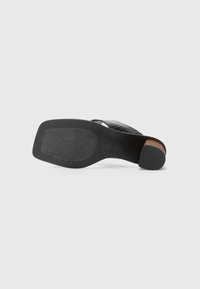 Sandale noire à talons avec une sangle en cuir texturé. Dispose d'une semelle en caoutchouc robuste, d'un talon en bois et d'un dessous lisse et plat pour une meilleure adhérence.