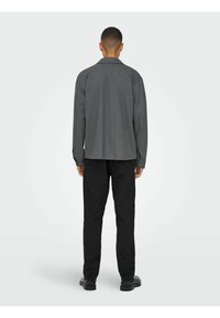 Graues Hemd mit entspannter Passform, langen Ärmeln und Knopfmanschetten, kombiniert mit schwarzen Hosen und schwarzen Schuhen. Glatte Textur, keine sichtbaren Muster.