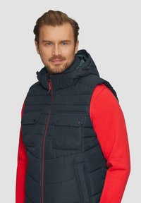 Gestepptes navyblaues Weste mit Kapuze, ausgestattet mit zwei Brusttaschen, roten Reißverschlussakzenten und gestepptem Muster. Getragen über einem roten Langarmshirt.