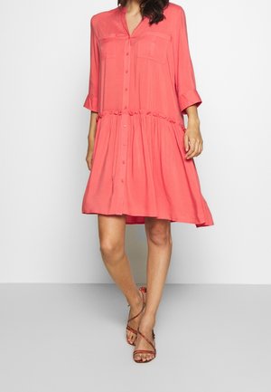 Frau trägt ein korallenfarbenes, knöpfbares Kleid mit geraffter Taille und dreiviertellangen Ärmeln, kombiniert mit hochhackigen Sandalen mit Riemchen.