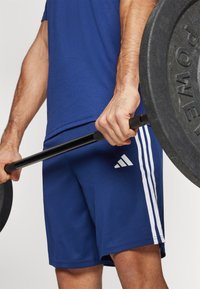 Shorts navy avec des rayures blanches, en tissu mélangé polyester, avec un logo Adidas. Personne tenant une barre de poids noire avec une poignée texturée.