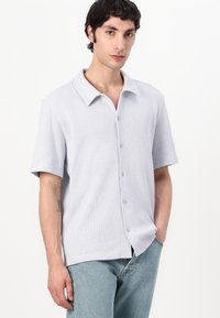 Camicia a maniche corte con colletto di colore grigio chiaro, caratterizzata da un motivo a maglia testurizzata e chiusura con bottoni. Abbinata a jeans in denim blu chiaro.