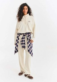 Béžový fleecový pulovr s krátkým zipem a logem, doplněný krémovými kalhotami. Modrá kostkovaná flanelová košile uvázaná v pase a hnědé tenisky.