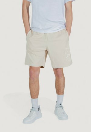 Pantaloni corti beige con tasche laterali, cintura elastica e logo "EA" bianco sulla parte inferiore della gamba destra. Indossati con una maglietta bianca e sneakers.