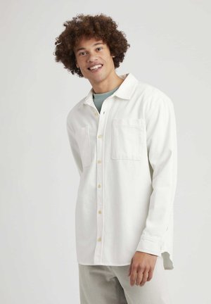 Chemise blanche à manches longues avec un col, deux poches poitrine, une fermeture à boutons sur le devant et une texture douce, portée par un modèle.