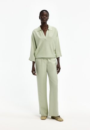 Frau trägt hellgrüne, langärmlige Bluse mit Kragen und passende, weite Hosen, steht und trägt flache, gemusterte Slip-On-Schuhe.