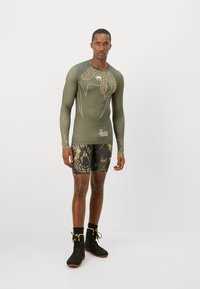 Camicia a maniche lunghe color oliva con design a foglia, abbinata a pantaloni corti neri con motivo. Scarpe sportive nere con dettagli gialli completano l'outfit.