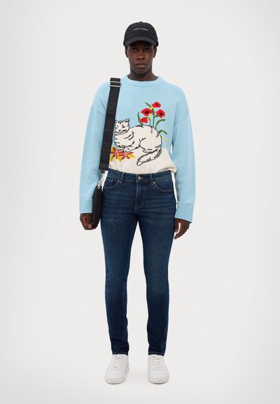 Light blue sweater with cat and flower design, dark jeans, and white sneakers. Accessory: black crossbody bag. Casual outfit.