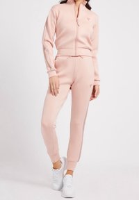 Světlý růžový teplákový komplet vyrobený z jemného materiálu. Zipová bunda s vysokým límcem a logem, doplněná o úzké kalhoty s bočními akcenty. Bílé tenisky.