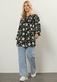 Tunika nera floreale con fiori bianchi, maniche lunghe a sbuffo e chiusura con cravatta, abbinata a jeans a gamba larga blu chiaro e sneakers bianche.