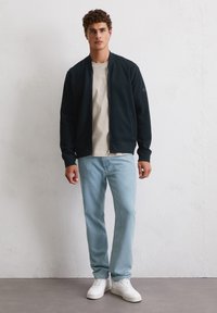 Black bomber jacket with ribbed cuffs and hem, worn over a light beige t-shirt, paired with light blue jeans and white sneakers.