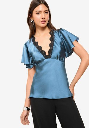 Top blu satin con maniche a sbuffo e scollatura a V profonda rifinita in pizzo nero. Texture morbida; indossato con pantaloni neri e un bracciale con dettagli argentati.