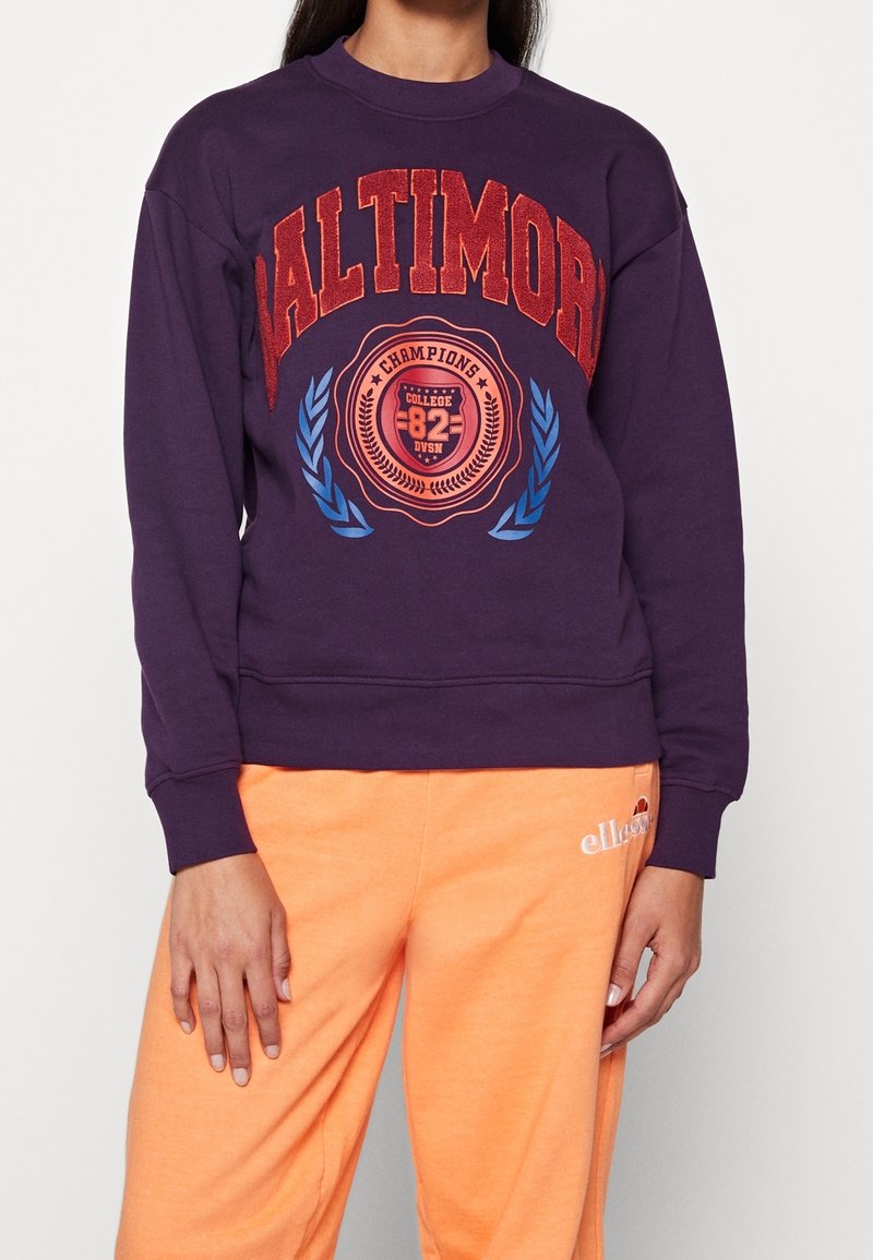 Personne portant un sweat-shirt violet avec le texte « Baltimore Champions College 82 » et des lauriers bleus, associé à un pantalon de survêtement orange.
