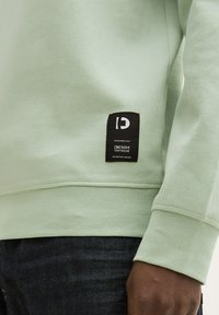 Sudadera verde claro hecha de tela texturizada con un cuello ajustado. Presenta una etiqueta de marca negra en el lado inferior.