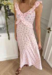 Femme portant une robe fleurie rose sans manches avec un col volanté, debout dans une cuisine près d'un plan de travail blanc et d'une porte.