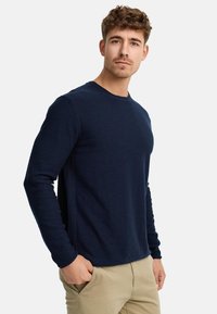 Maglietta navy a maniche lunghe con colletto a girocollo in un tessuto a trama, dotata di polsini a coste e orlo dritto, abbinata a pantaloni beige chiaro.