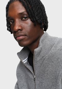 Close-up van een persoon met ingevlochten haar, die een grijze ritsvest draagt over een zwart shirt, tegen een effen achtergrond.