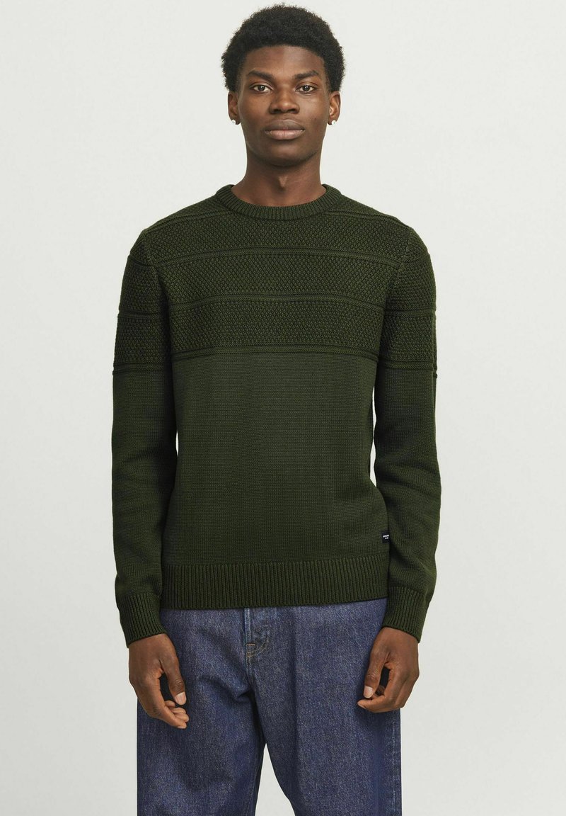 Pull en tricot vert foncé avec des motifs texturés, col rond, et ourlet et poignets côtelés. Associe avec un jean bleu.