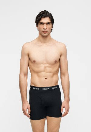 Homme torse nu aux cheveux courts et foncés portant un boxer noir Hollister, debout face à un fond clair uni.