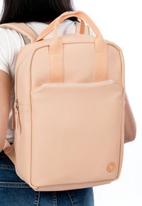 Persikanvärinen reppu, jossa on sileä, mattapintainen pinta. Siinä on kaksi yläkahvaa, etutasku vetoketjulla ja pyöreä logokoriste.