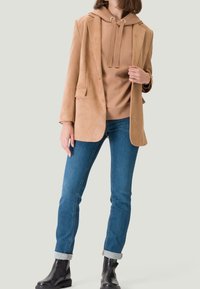 Beige suède blazer gedragen over een beige hoodie met trekkoorden, gecombineerd met blauwe spijkerbroek en zwarte chunky laarzen. Eenvoudig ontwerp met minimale accenten.