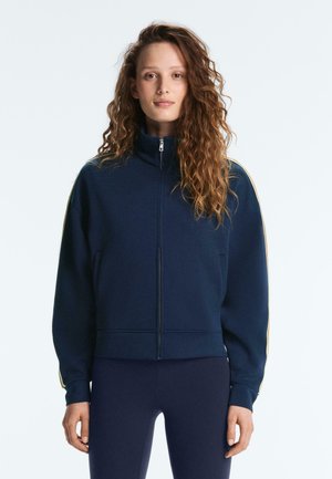 Hosszú, göndör hajú nő, tengerészkék cipzáros kabátot visel, sárga csíkokkal az ujján, valamint hozzá illő tengerészkék leggingset.