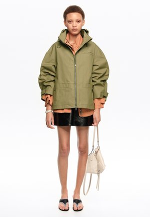 Persona joven con una chaqueta verde oliva de gran tamaño, camiseta naranja, falda negra, sandalias negras, sosteniendo un pequeño bolso beige.