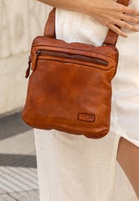 Hnědá kožená crossbody taška s zipovou přední kapsou, hladkým povrchem a nastavitelným popruhem. Logo značky zobrazeno na boku.