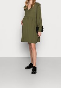 Olivi vihreä äitiysmekko, jossa on V-pääntie, kerrostettu vyötärö, pitkät hihat ja hillitty olkapääyksityiskohta, yhdistetty mustiin lakattuihin kenkiin.