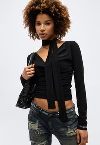 Νεαρή γυναίκα με κυματιστά μαλλιά, φοράει μαύρη μπλούζα με μακριά μανίκια και δετή λαιμόκοψη, σκισμένο σκούρο τζιν και κρατάει μια μαύρη γυαλιστερή τσάντα ώμου.