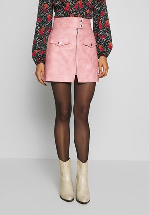 Mujer con minifalda rosa de talle alto con cremallera y bolsillos delanteros, medias negras transparentes, blusa floral y botines blancos hasta el tobillo.