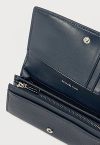 Πορτοφόλι από γαλάζιο δέρμα με υφή. Διαθέτει τσέπη με φερμουάρ, θέσεις για κάρτες και κλείσιμο με πατιλέτα. Λογότυπο χαραγμένο στο εσωτερικό.