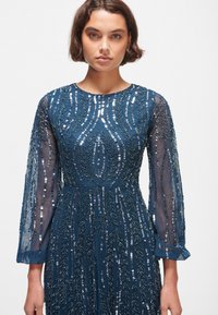 Robe bleu marine avec de longues manches transparentes, embellies de motifs de feuilles en sequins ; taille cintrée et jupe fluide. Design élégant avec des détails complexes.