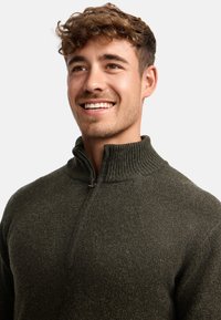 Joven con cabello rizado sonriendo, vistiendo un suéter de punto verde oscuro con media cremallera y cuello acanalado, contra un fondo sencillo.