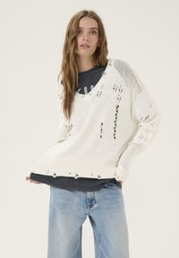 Chunky witte trui met een gestructureerde brei, grote v-hals, versleten details en ribbelrand, gelaagd over een grijze t-shirt en gecombineerd met blauwe spijkerbroek.