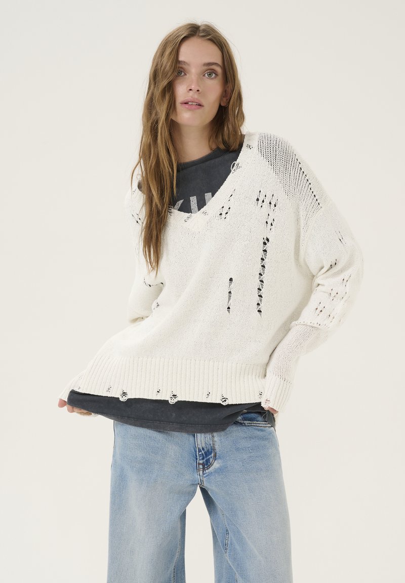 Chunky witte trui met een gestructureerde brei, grote v-hals, versleten details en ribbelrand, gelaagd over een grijze t-shirt en gecombineerd met blauwe spijkerbroek.