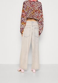 Henkilö, jolla on päällään vaaleansiniset löysät farkut, vaaleanpunaiset korkopohjaiset kengät ja värikäs raidallinen lyhyt neule, seisoo harmaata taustaa vasten.