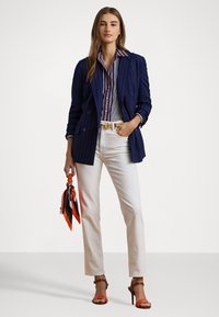 Blazer doppiopetto a righe blu navy, pantaloni bianchi a gamba dritta, camicia a righe multicolore, accessoriate con una cintura e una sciarpa arancione.