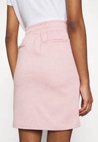 Person wearing a light pink elastic waist skirt and white top, shown from the back from mid-torso to mid-thigh against a white background.