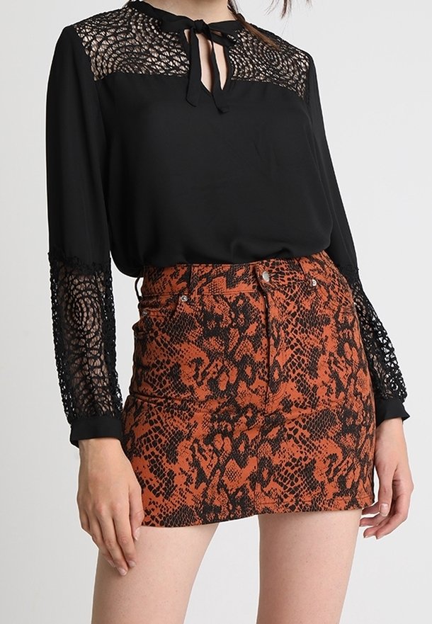 Blusa nera con maniche in pizzo e fiocco al collo, abbinata a una gonna aderente arancione e nera con motivo a serpente, creando un look a contrasto.