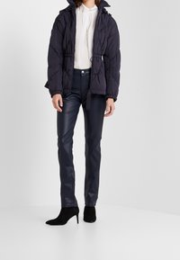 Marineblauer Pufferjacke mit Kordelzug in der Taille, kombiniert mit figurbetonten marineblauen Kunstlederhosen und schwarzen hochhackigen Stiefeln. Glatte Textur insgesamt.