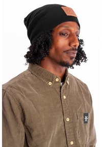 Black knit beanie with a tan leather patch. Model wears a brown corduroy shirt with button details and a left chest pocket logo.