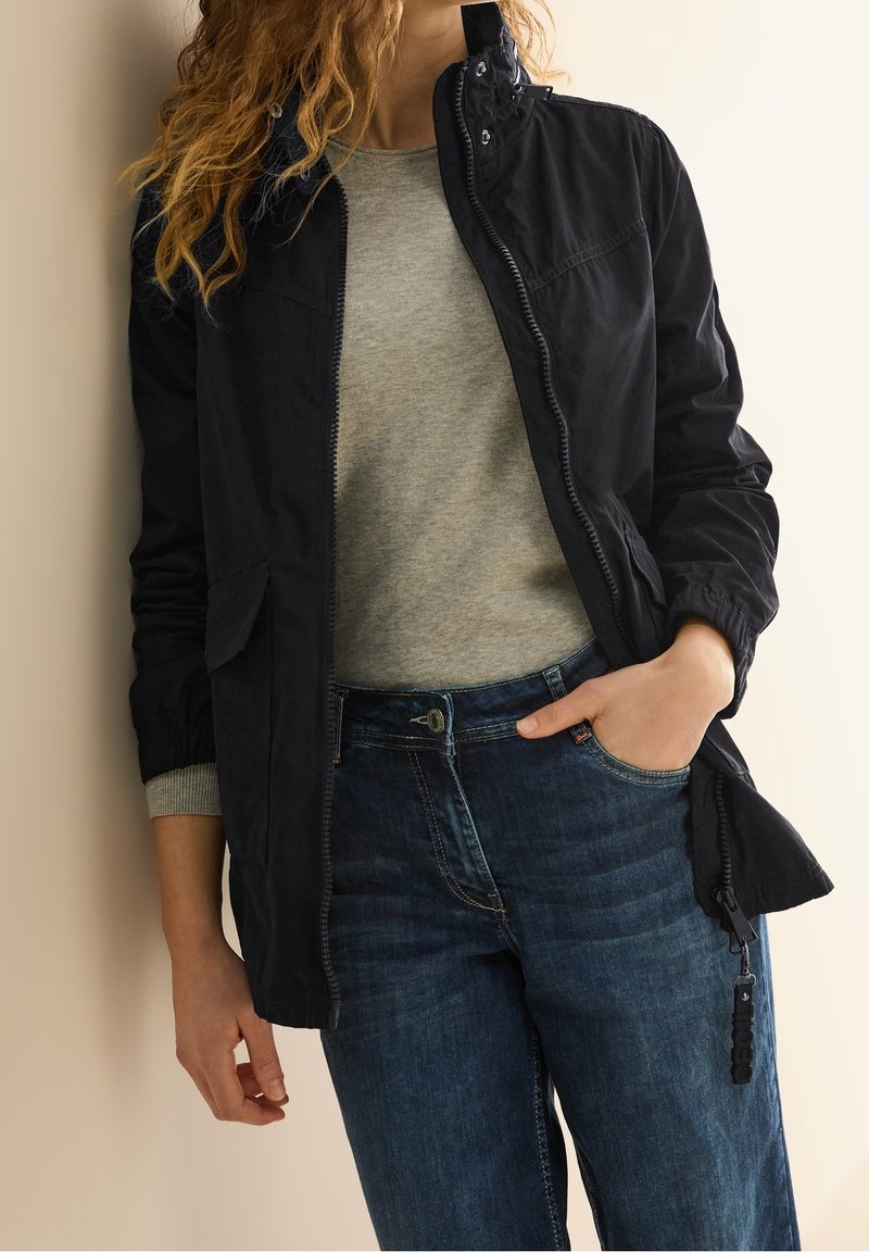 Black zip-up jacket with high collar, side pockets, and elastic cuffs, paired with a gray top and dark blue jeans. Smooth texture.