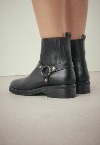 Botas de tobillo de cuero negro con un eje acanalado, detalles en hardware dorado y un tacón grueso. Presentan una correa con un detalle de hebilla redonda.