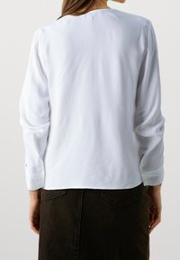 Vit långärmad blus med en slät yta och rund halsringning. Har knäppta manschetter och en avslappnad passform bak.