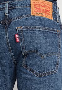 Mörkblå denimjeans med en texturerad yta. Funktioner inkluderar en läderlogga, röd etikett och gulddetaljer på fickorna.