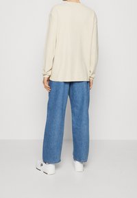 Beżowa bluzka z długim rękawem o luźnym kroju, w połączeniu z szerokimi niebieskimi dżinsami. Białe sneakersy na platformie z subtelnymi kolorowymi akcentami.