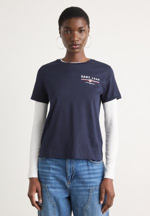 T-shirt blu navy con collo rotondo, maniche corte e grafica "GANT 1949"; indossata sopra una camicia bianca a maniche lunghe.